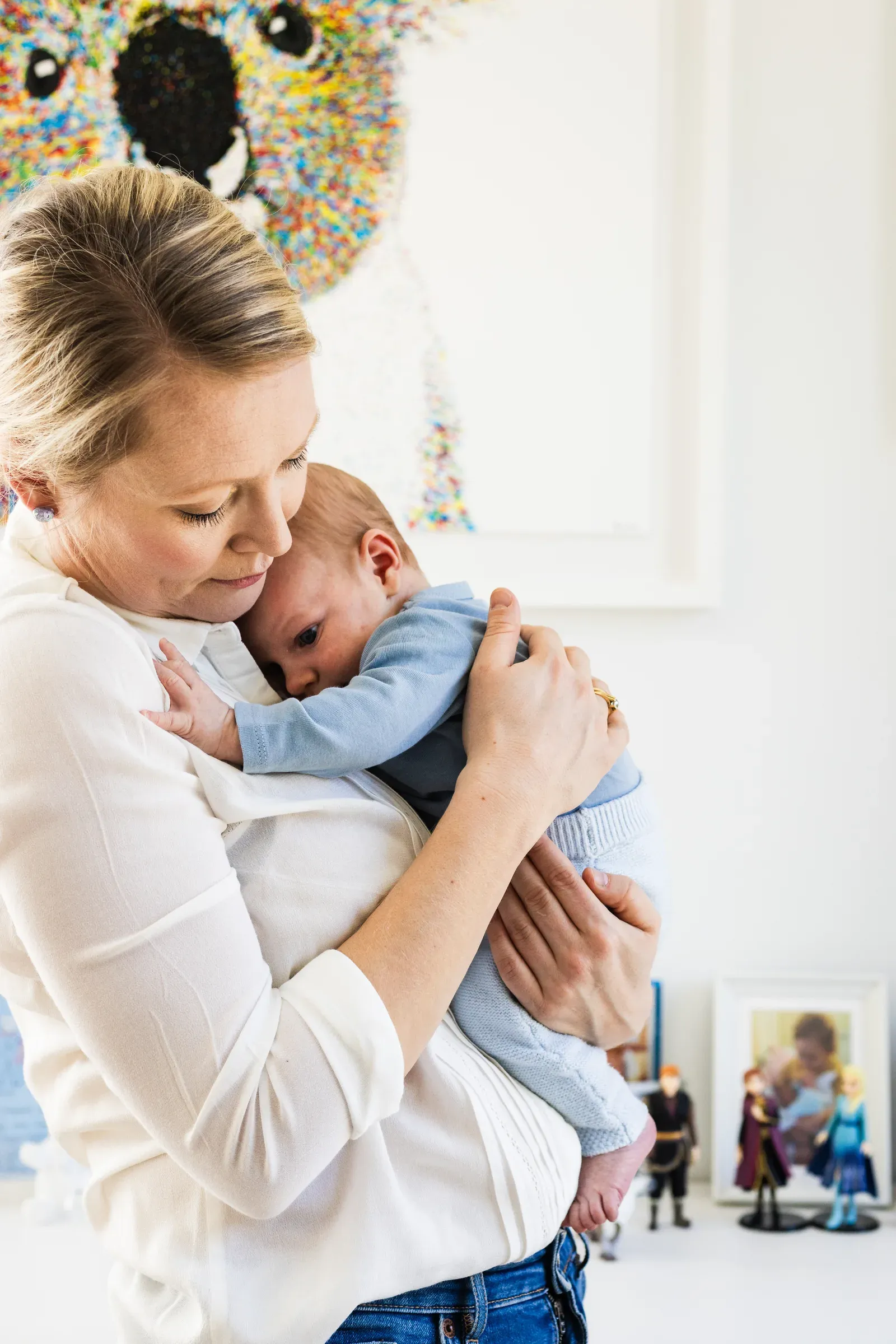 Mags cradling her baby, eye to eye, in warm window light.